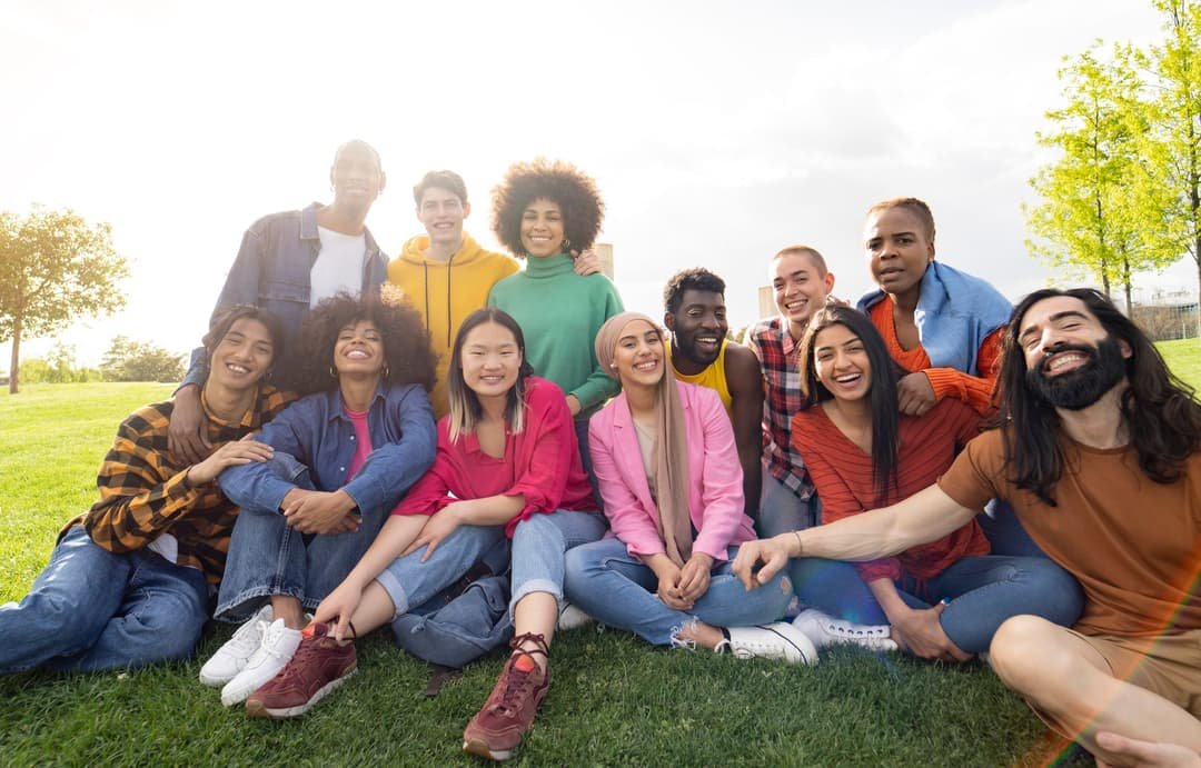 A large group of friends from many different backgrounds sitting together on the grass in a park, smiling.